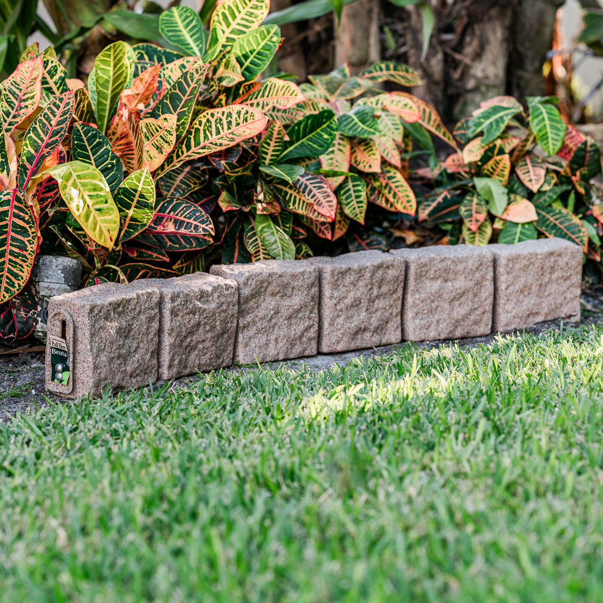 Row of gray PolyRock edging blocks bordering a garden bed with vibrant croton leaves and a grassy foreground